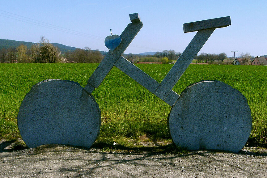 Sehr großes Fahrrad aus Stein am Wegesrand. Im Hintergrund große grüne Weide.