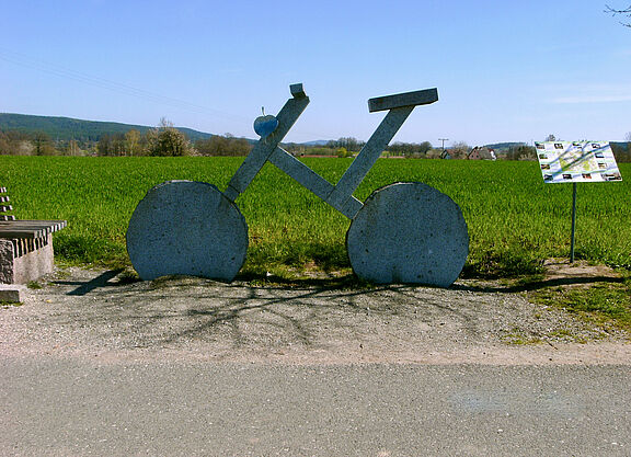 Ein übergroßes Fahrrad aus Stein steht am Wegesrand vor einer grünen Wiese.