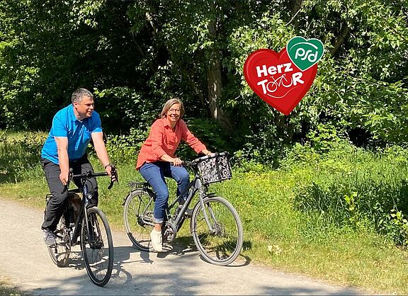 Eine Radfahrerin und ein Radfahrer radeln auf einem naturbelassenem Weg durch eine baumreiche Gegend. Ein rotes Herz mit der Inschrift HerzTour, wobei das ou durch ein Fahrrad symbolisiert wird.