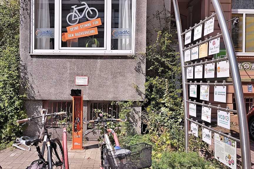 Hinter roten Fahrradbügeln steht eine orangefarbende Fahrradreparatursäule. Darüber ist das Fenster der ADFC-Geschäftsstelle zu sehen mit der Beschriftung "Deine Stimme für den Radverkehr" unter einem weißem Fahrrad.