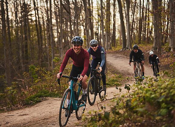 Mit Gravelbikes abseits des Asphalts unterwegs. Eine Gruppe von Frauen und Männern fahren mit Gravelbikes hintereinander auf einem Waldweg.
